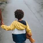 From above back view of crop anonymous ethnic father holding boy with toy bear by hand on asphalt roadway