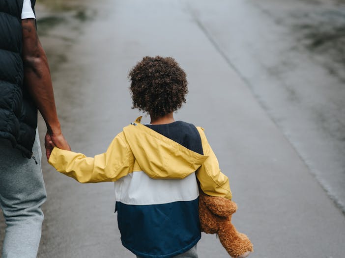 From above back view of crop anonymous ethnic father holding boy with toy bear by hand on asphalt roadway